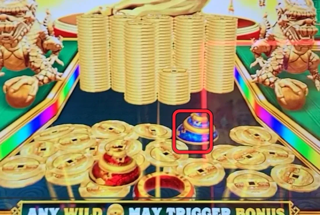 The scouting read, ringed: a blue-and-gold progressive GEM parked right near the FRONT EDGE of the tray with the tall coin stacks built up behind it — exactly the overlooked 'lock' play. A previous player built the prize equity and walked without collecting it. Read the tray, not the reels; gems can look like ordinary prize balls (note the red ball beside it), so inspect the front edge carefully.