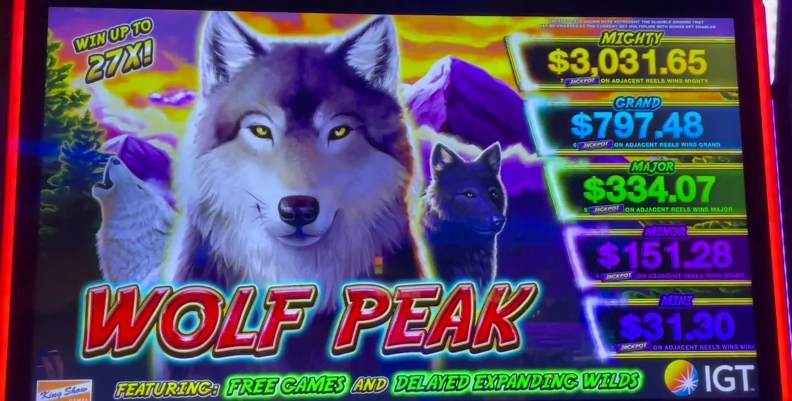 Know it on sight: the WOLF PEAK wordmark and wolf face, 'WIN UP TO 27X!', the Mighty/Grand/Major/Minor/Mini jackpot ladder, and the on-glass tagline 'FEATURING: FREE GAMES AND DELAYED EXPANDING WILDS' — the mechanic that makes this an advantage play is named right on the glass.